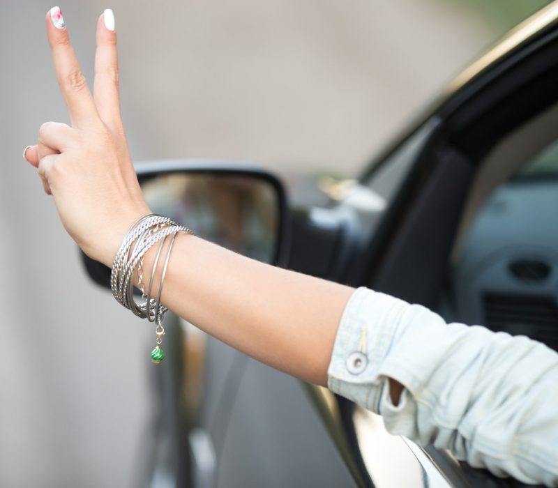 Beautiful young woman in casual denim jacket riding modern cabriolet car, showing victory gesture through the window, close up of hand with cute fashion bracelet, back view